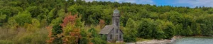 Verweerde houten vuurtoren met een klein huisje aan de oever, omgeven door dicht bos met herfstkleuren in Michigan