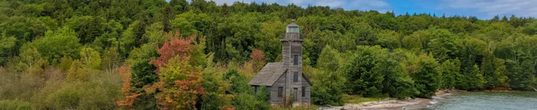 Verweerde houten vuurtoren met een klein huisje aan de oever, omgeven door dicht bos met herfstkleuren in Michigan