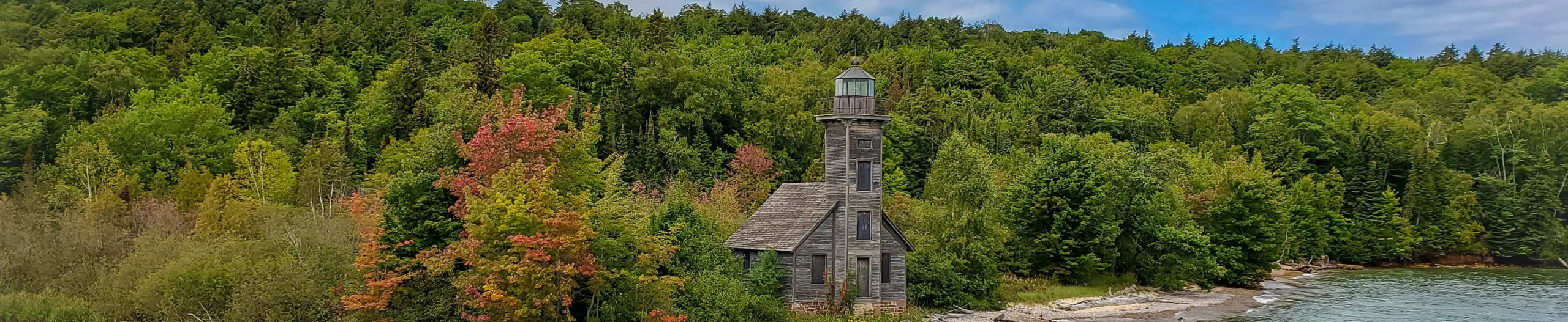 Verweerde houten vuurtoren met een klein huisje aan de oever, omgeven door dicht bos met herfstkleuren in Michigan