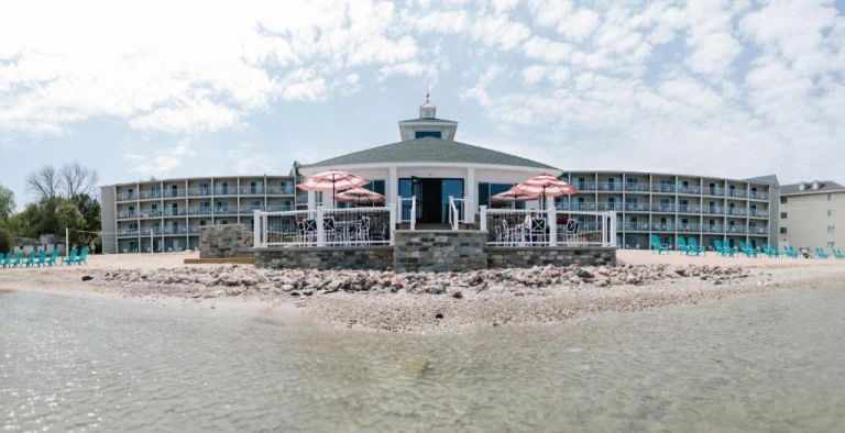 Wit hotelgebouw met balkons en een terras met roze parasols direct aan het strand van een meer in Michigan