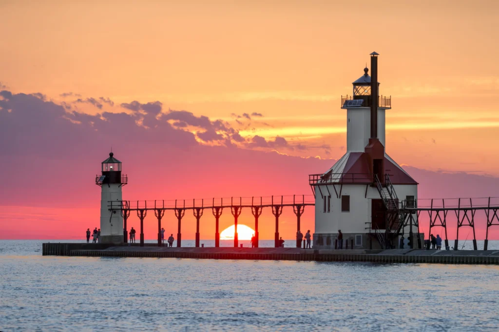 Twee witte vuurtorens op een pier met mensen die uitkijken over het meer terwijl de zon onder het water zakt in Michigan