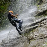Een man met oranje helm abseilt langs een rotswand terwijl water van een waterval langs hem naar beneden stroomt