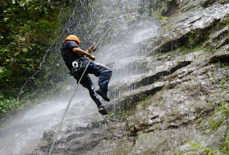 Een man met oranje helm abseilt langs een rotswand terwijl water van een waterval langs hem naar beneden stroomt
