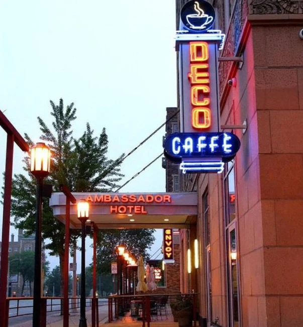 Kleurrijke neonreclames van Deco Caffe en het Ambassador Hotel boven een overdekte stoep bij het vallen van de avond