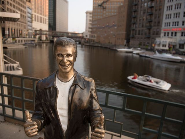 Bronzen standbeeld van de Fonz met opgestoken duimen aan de Milwaukee Riverwalk met een boot en gebouwen op de achtergrond