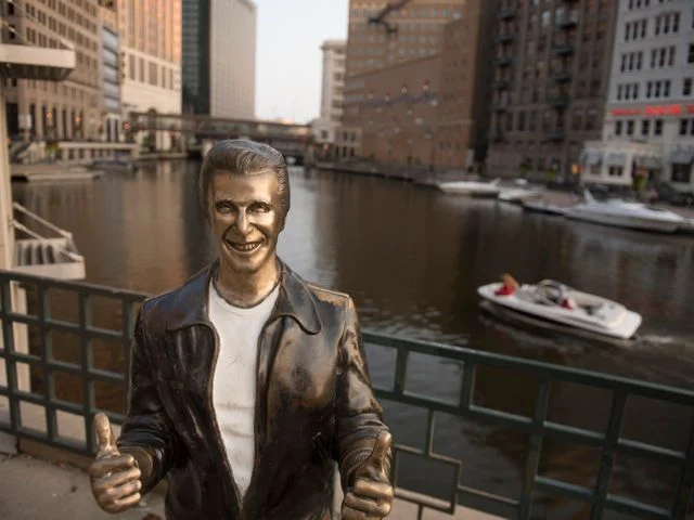 Bronzen standbeeld van de tv figuur The Fonz met beide duimen omhoog voor een reling langs de rivier in Milwaukee met een boot op de achtergrond