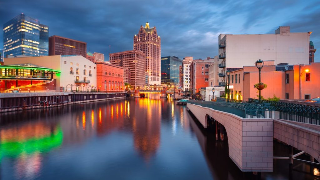 Verlichte gebouwen langs de Milwaukee River die weerspiegelen in het water bij schemering, met een voetgangersbrug op de voorgrond