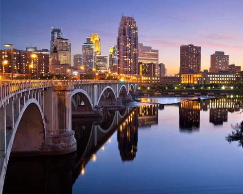 Verlichte wolkenkrabbers van Minneapolis weerspiegeld in het water naast een stenen boogbrug bij schemering