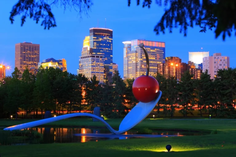 Het kunstwerk Spoonbridge and Cherry in de sculpture garden van Minneapolis met de verlichte skyline op de achtergrond bij schemering