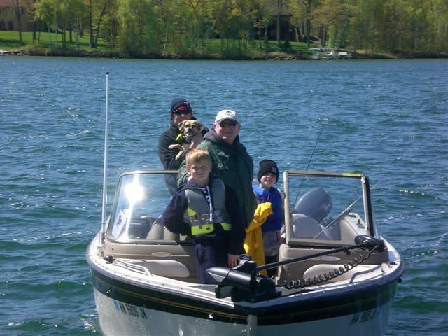 Twee mannen, twee kinderen en een hond in een visboot op een meer, met bomen op de achtergrond