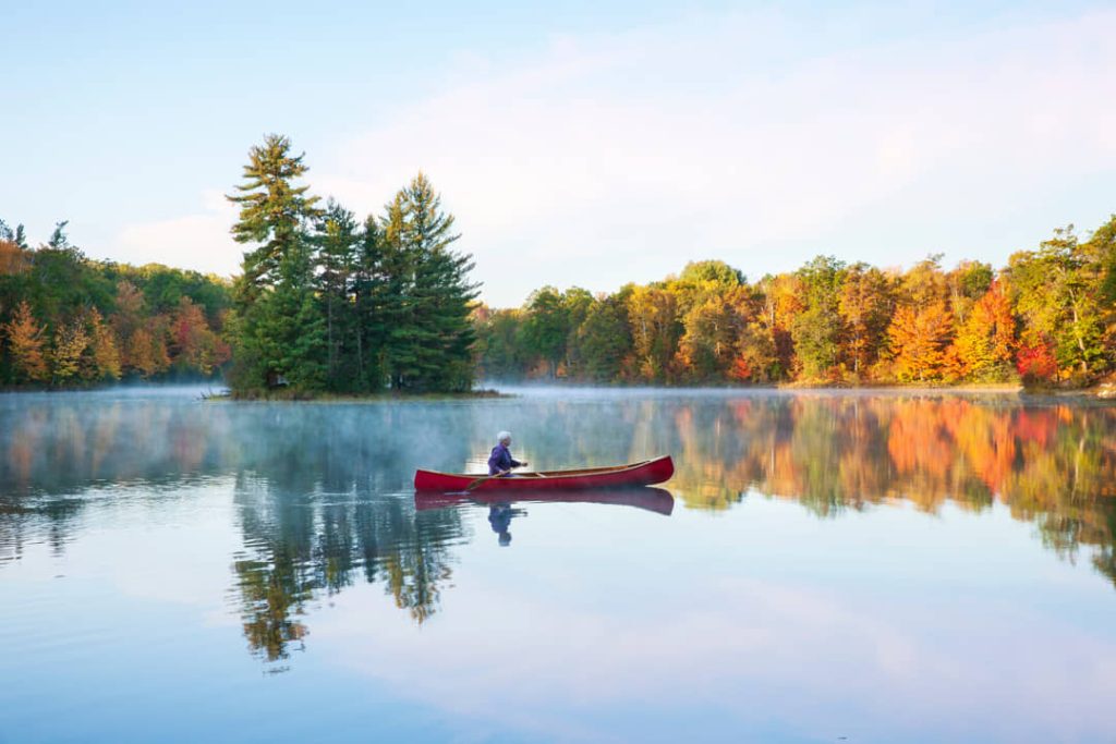 Een persoon peddelt in een rode kano over een rustig meer in Minnesota, omringd door bomen in herfstkleuren en ochtendnevel boven het water