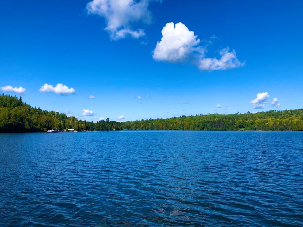 Rustig blauw meer omringd door bos, met enkele huisjes aan de overkant onder een wolkenlucht