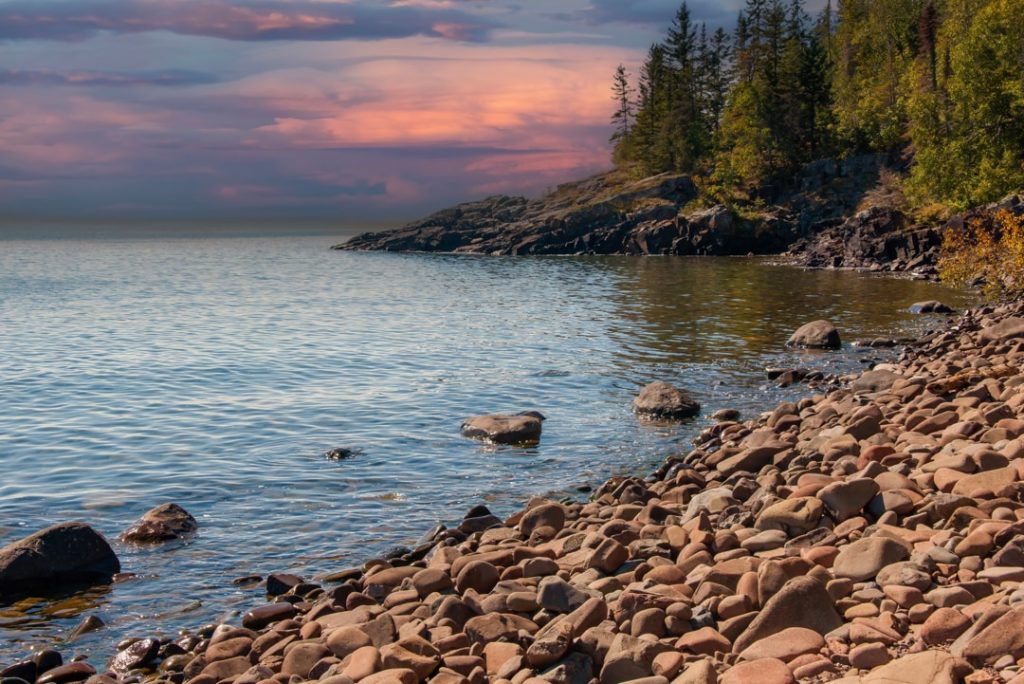 Rond kiezelstrand langs Lake Superior met zacht water, dennenbomen en een gekleurde avondlucht op de achtergrond