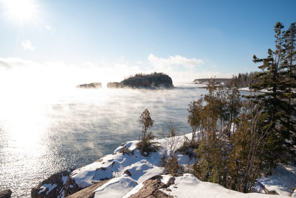 Opstijgende mist boven het water van Lake Superior in de winter, met besneeuwde rotsen en dennenbomen op de voorgrond
