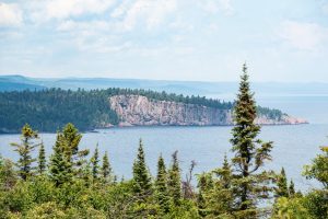 Uitzicht tussen dennentakken door op een rotsachtige kaap die ver het water van Lake Superior in steekt