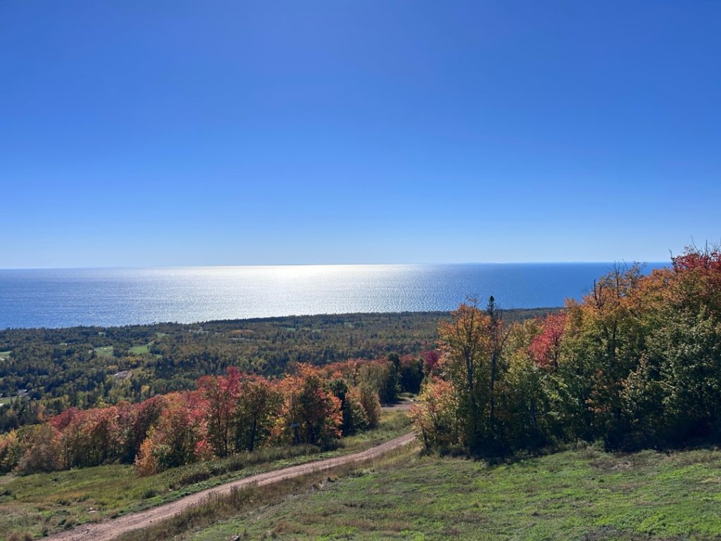 Uitzicht vanaf een heuvel met een zandweg over bos in herfstkleuren, met Lake Superior glinsterend op de achtergrond