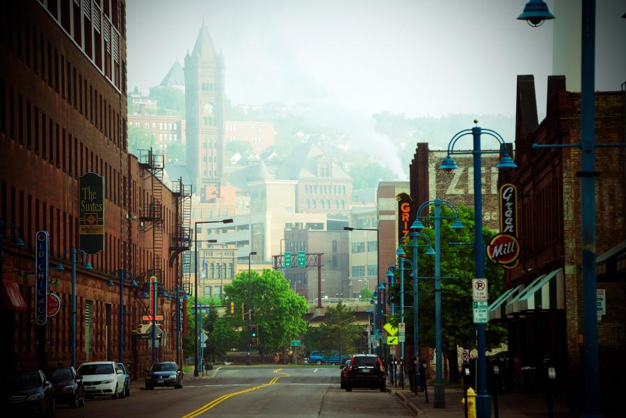 Uitzicht over een historische straat in Duluth, Minnesota, met bakstenen gebouwen, straatlantaarns en de skyline van de stad in de mist op de achtergrond