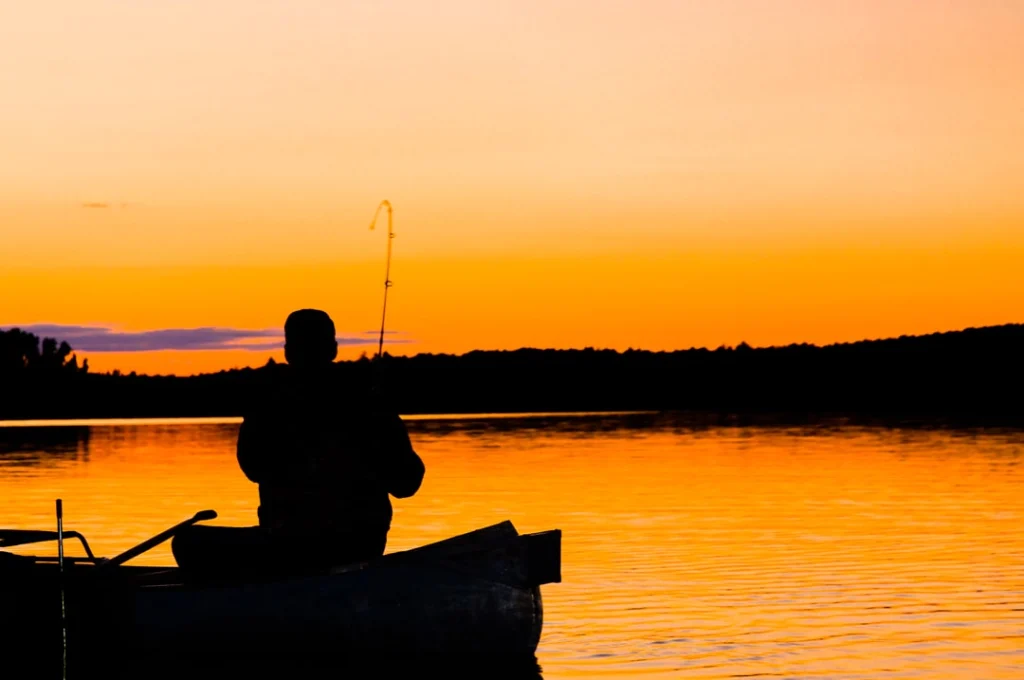 Silhouet van een visser met hengel in een boot op een meer tijdens een oranje zonsondergang