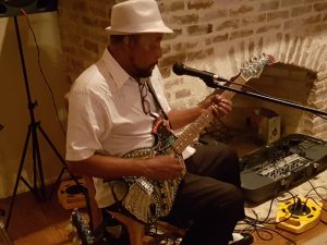 Man met witte hoed speelt elektrische gitaar bij een microfoon in een kleine club met bakstenen muren en versterkerapparatuur