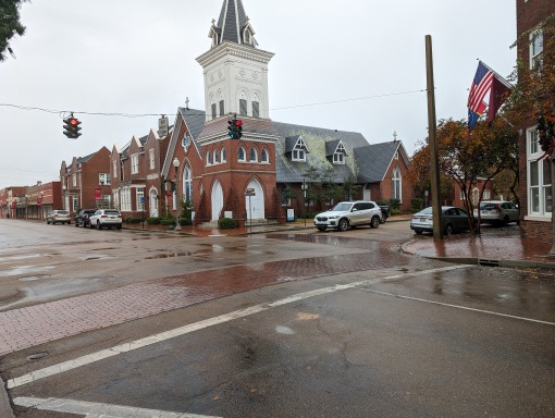 Rood bakstenen kerk met een witte torenspits op een straathoek met verkeerslichten en geparkeerde auto's bij regenachtig weer in Mississippi