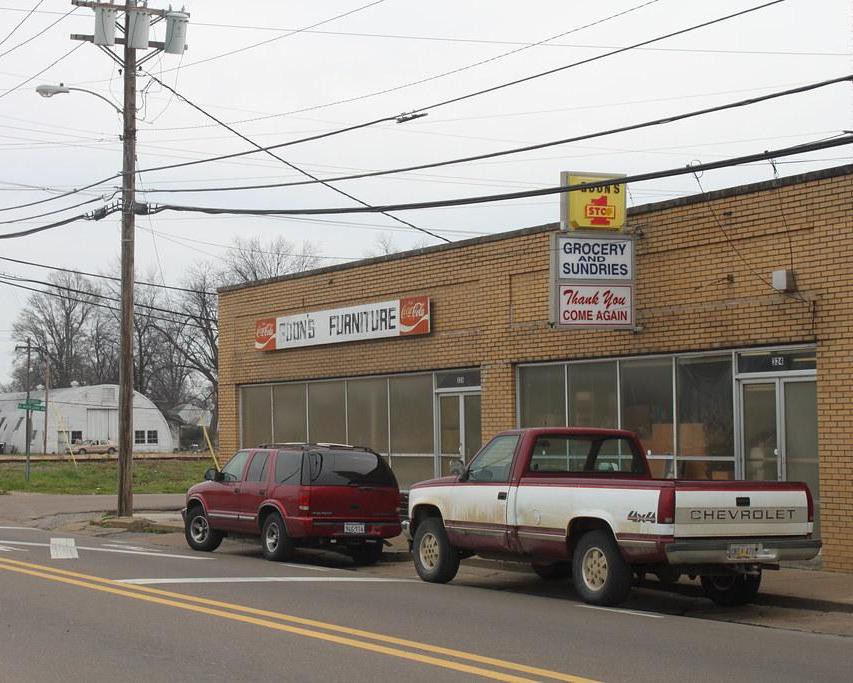 Bakstenen winkelpand met de opschriften Coon's Furniture en Grocery and Sundries met twee oldtimer pick-up trucks ervoor in Mississippi