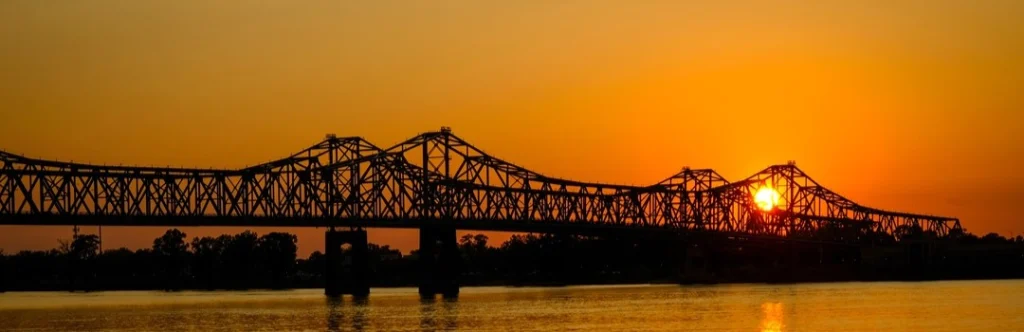 Stalen vakwerkbrug in silhouet over de brede Mississippi rivier met een felle oranje zonsondergang op de achtergrond
