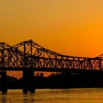 Stalen vakwerkbrug in silhouet over de brede Mississippi rivier met een felle oranje zonsondergang op de achtergrond