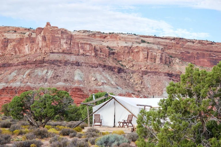 Een witte glampingtent met terrasje staat aan de voet van een hoge rode rotswand bij Moab in Utah