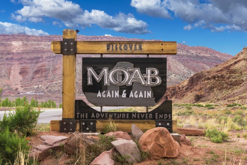 Houten bord met de tekst Discover Moab Again and Again voor een rode rotswand langs de weg