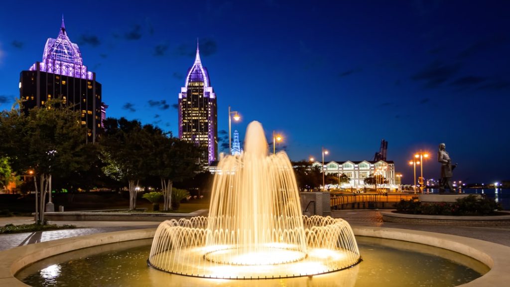 Hoge klaterende fontein in een park bij nacht met de paars verlichte wolkenkrabbers van Mobile in Alabama op de achtergrond
