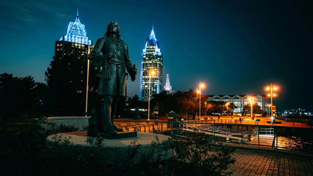 Bronzen standbeeld van een historische figuur op de kade met de verlichte wolkenkrabbers van Mobile in Alabama bij nacht