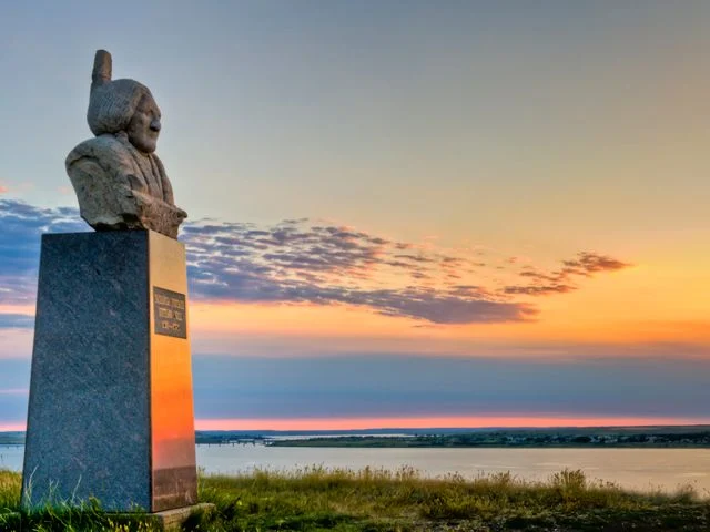 Stenen buste van een inheemse Amerikaanse leider staat op een sokkel met uitzicht over het water bij zonsondergang