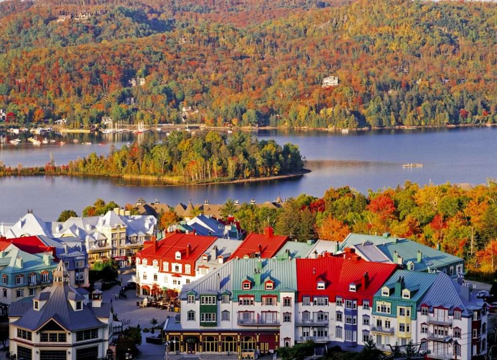 Luchtfoto van de kleurrijke daken van Mont-Tremblant naast een meer met eilandje, omringd door bergen vol herfstkleuren