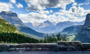 Uitzicht over een groen dal met steile bergtoppen gezien vanaf een uitkijkpunt met een stenen muur in Glacier National Park