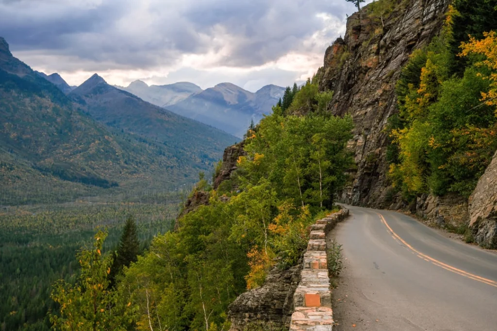 Kronkelende bergweg langs een rotswand met kleurrijke bomen en uitzicht op besneeuwde bergtoppen in Glacier National Park
