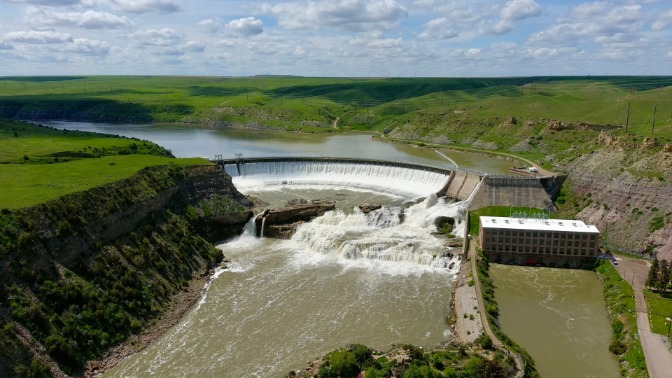 Luchtfoto van een stuwdam waarover water valt in een rivier, omringd door glooiende groene heuvels en een waterkrachtcentrale