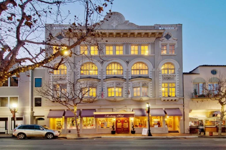 Verlicht historisch hotelgebouw met boogramen en luifels aan een straat met bomen en een geparkeerde auto in Monterey