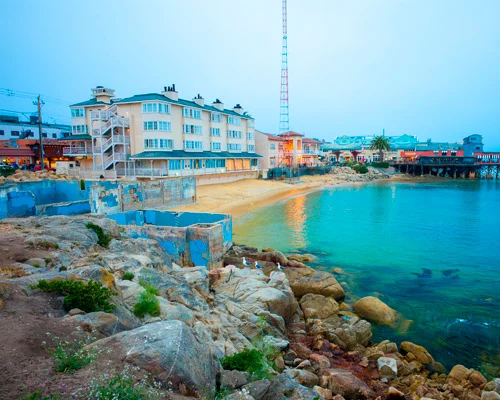 Rotsachtige kustlijn met een klein strand en hotelgebouwen aan het water bij schemering in Monterey
