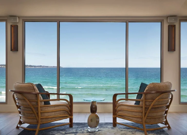 Twee rieten loungestoelen staan voor een groot raam met uitzicht op de oceaan in een hotelkamer bij Monterey
