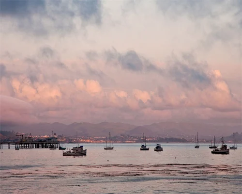Kleine vissersbootjes verspreid over kalm water met een roze wolkenlucht en heuvels op de achtergrond bij Monterey