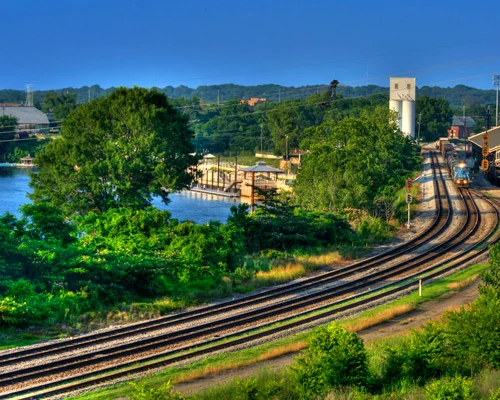Gebogen spoorrails langs een rivier met een graansilo en een trein op de achtergrond in het ochtendlicht bij Montgomery in Alabama