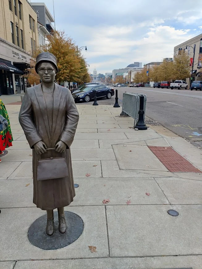 Bronzen standbeeld van een vrouw met een tas op een straathoek in het centrum van Montgomery in Alabama, met de koepel van het capitool in de verte