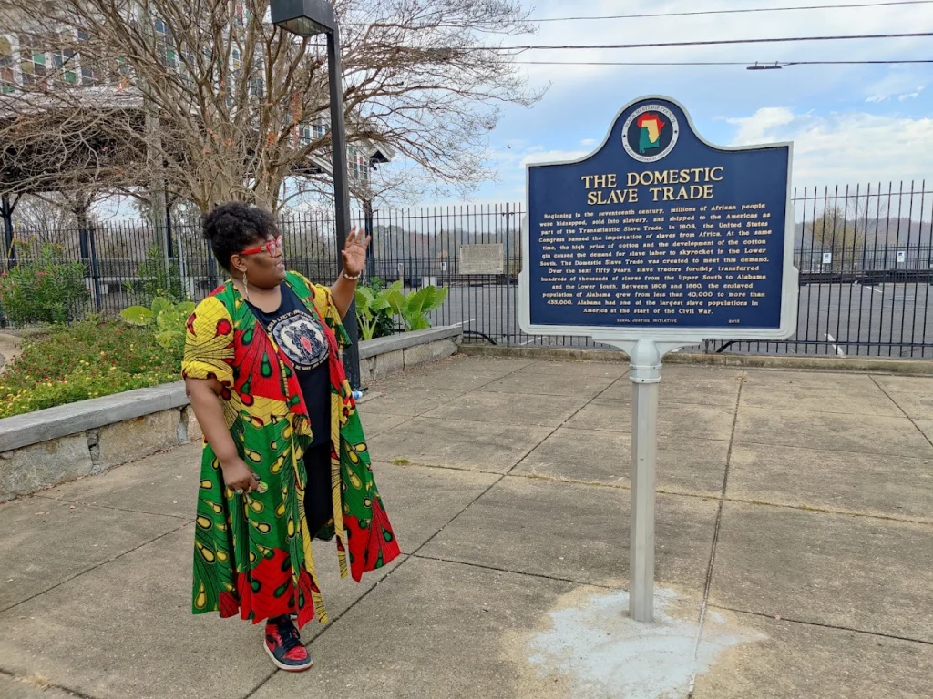 Vrouw in kleurrijke Afrikaanse kleding gebaart naar een historisch informatiebord over de binnenlandse slavenhandel in Montgomery in Alabama