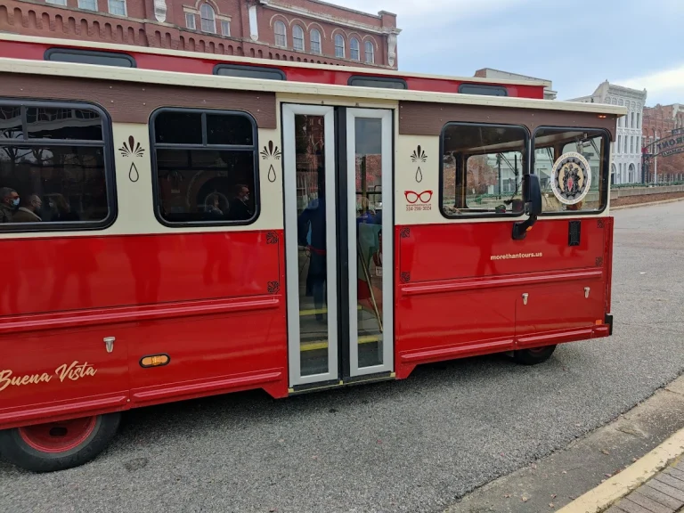 Een rode en crèmekleurige trolleybus met open deuren staat geparkeerd langs een straat in Montgomery, met historische bakstenen gebouwen op de achtergrond