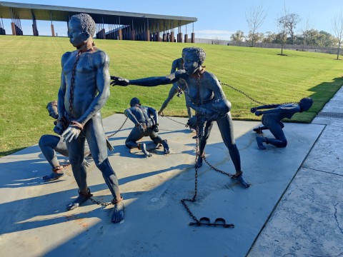 Bronzen standbeelden van tot slaaf gemaakte mensen in ketenen op een grasveld in Montgomery, met een modern gebouw op de achtergrond