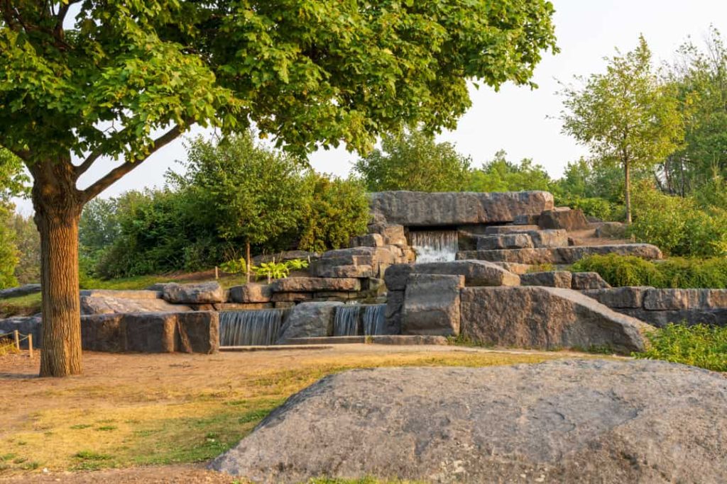 Kunstmatige waterval tussen grote rotsblokken in een groen park, met een grote boom op de voorgrond