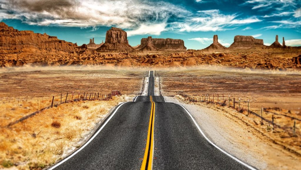 Rechte tweebaansweg loopt naar de horizon tussen droge velden met omheining, met de karakteristieke rode rotstorens van Monument Valley op de achtergrond