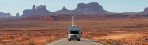 Een witte camper rijdt over een verlaten weg door de rode woestijn van Monument Valley, met markante rotsformaties op de achtergrond