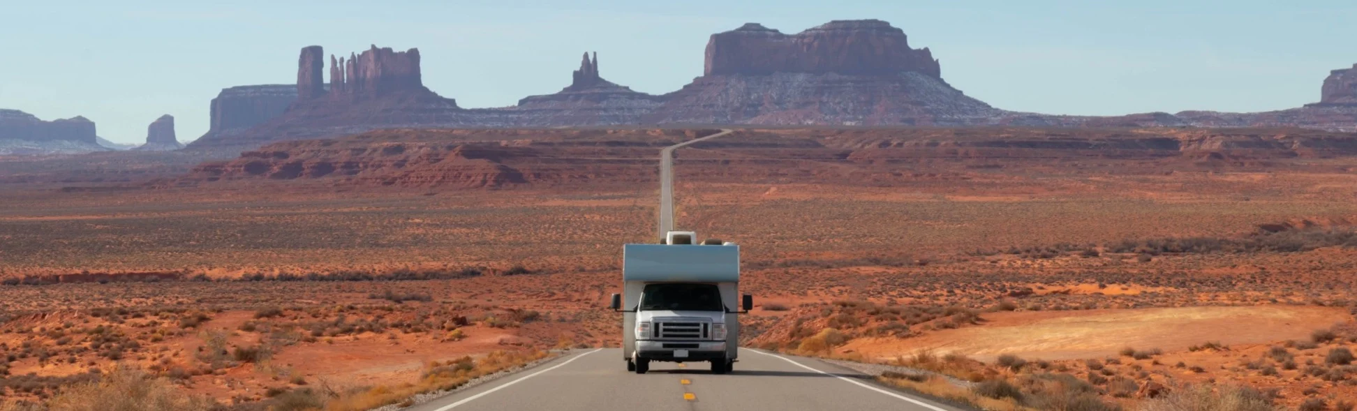 Een witte camper rijdt over een verlaten weg door de rode woestijn van Monument Valley, met markante rotsformaties op de achtergrond