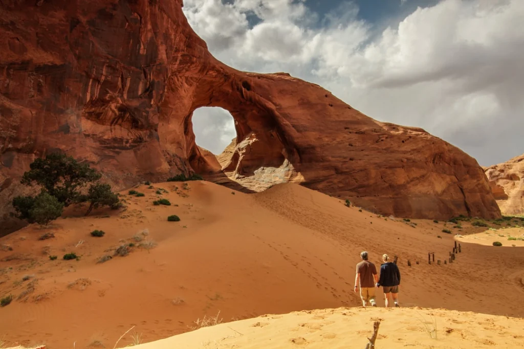 Twee wandelaars staan op roodkleurig zand en kijken naar een grote natuurlijke rotsboog in Monument Valley